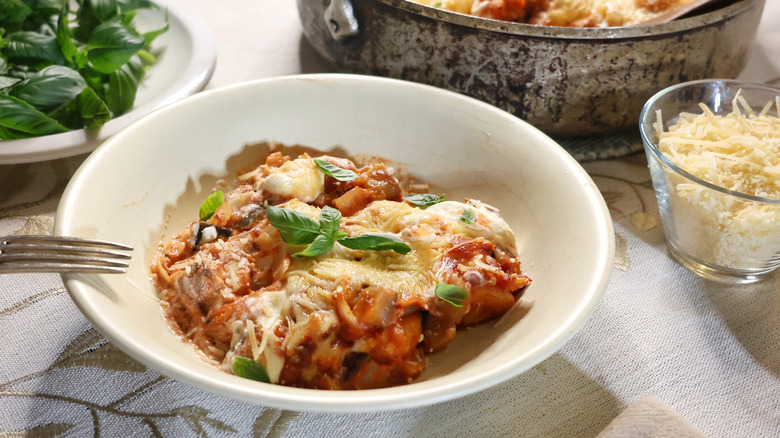 shallow bowl of gnocchi lasagna topped with fresh basil beside plate of basil, shredded parmesan cheese, and pan of baked gnocchi