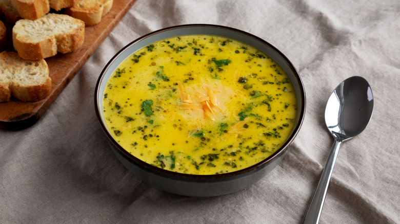 Bowl of broccoli Cheddar soup next to a spoon and slices of crusty baguette