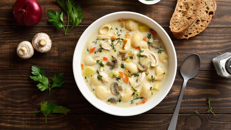 Bowl of creamy vegetable soup with pasta, next to a spoon and sliced bread