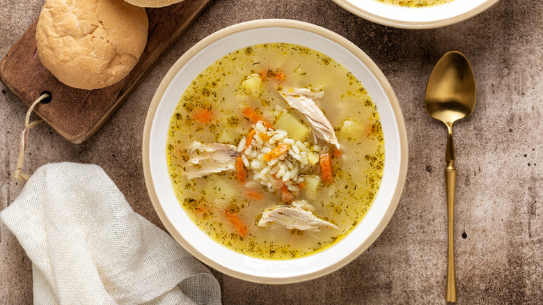 Bowl of chicken and rice soup next to a gold spoon and bread rolls
