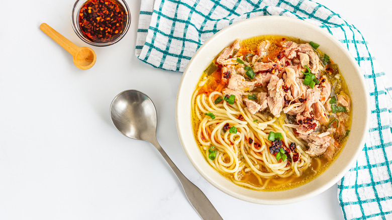 Bowl of chicken noodle soup next to a spoon and a small bowl of red pepper flakes