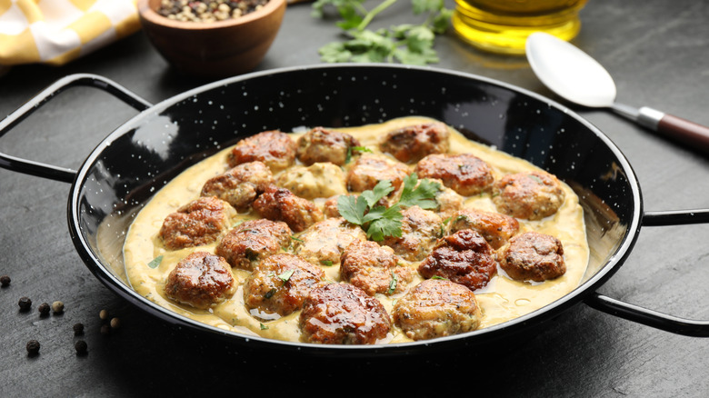 meatballs in cream gravy, cooked and served in a black-enameled paella pan, on a dark-colored tabletop with herbs, spices and oil visible to the rear