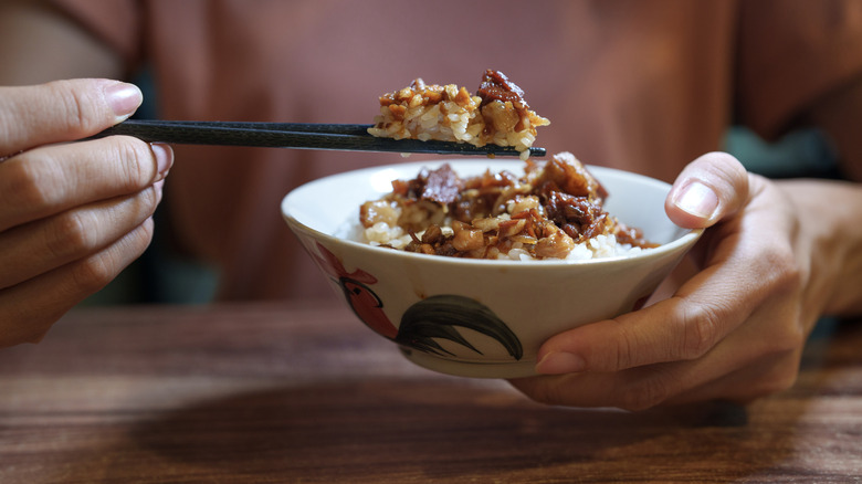 a bowl of braised pork rice held in a young woman's hand, with a mouthful of the dish held on her chopsticks in the other hand, over a wooden tabletop with her torso visible behind the bowl