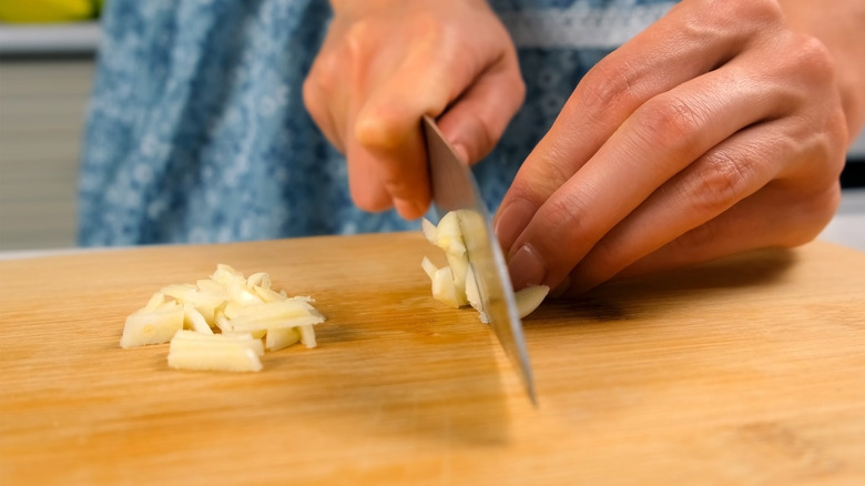 closeup of a woman's hands chopping fresh garlic with a wide-bladed knife on a wooden work surface, with her patterned blue dress (slightly old-fashioned looking) visible but out of focus behind her hands