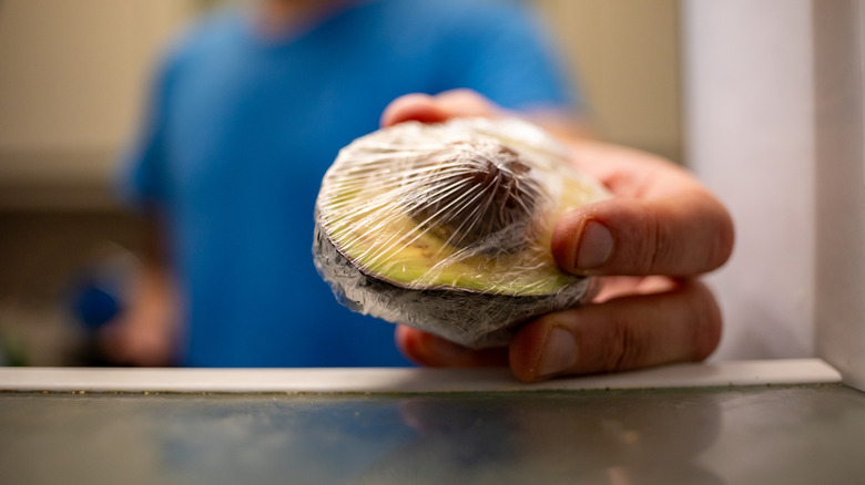 Man placing avocado with pit in plastic wrap in the refrigerator