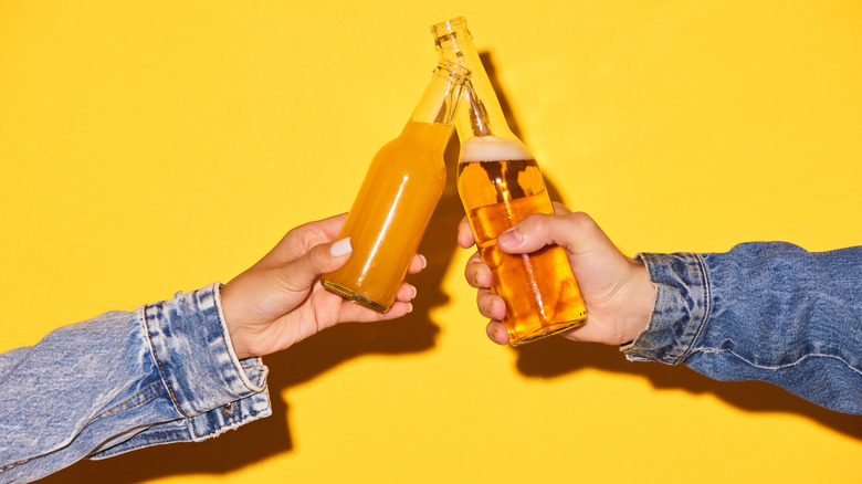 Two hands holding bottles of beer against yellow background and toasting