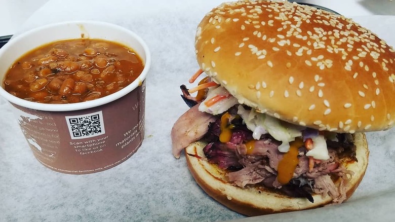Cardboard container of baked beans on wax paper next to a sandwich on a sesame seed bun bursting with chopped meat, coleslaw, and sauce