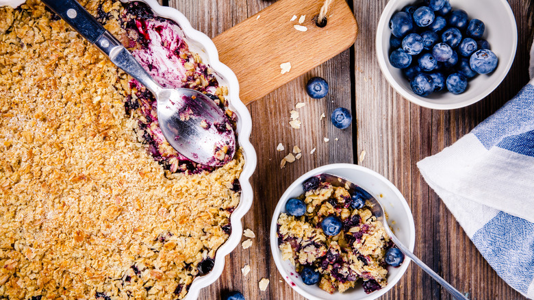 overhead shot of blueberry crisp with spoon in a large and small dish and some blueberries on the side on a wooden table