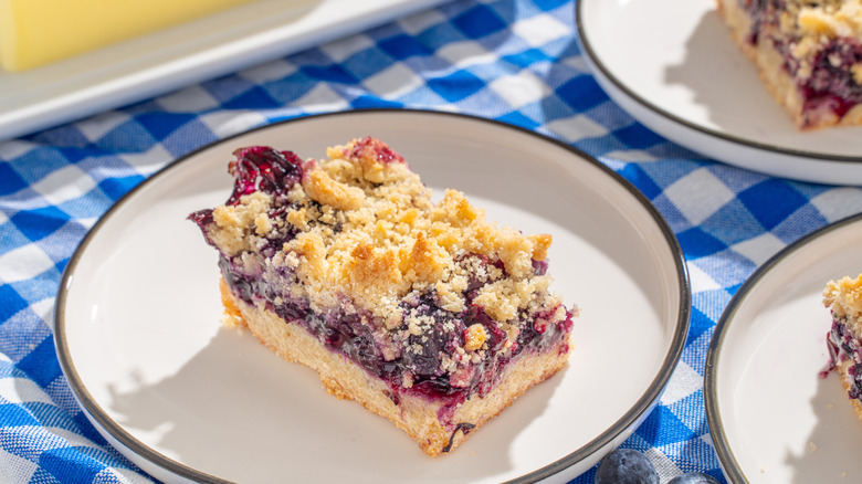 blueberry crumb bar serving on a plate with a blue checkered kitchen towel in the back and more plates around it