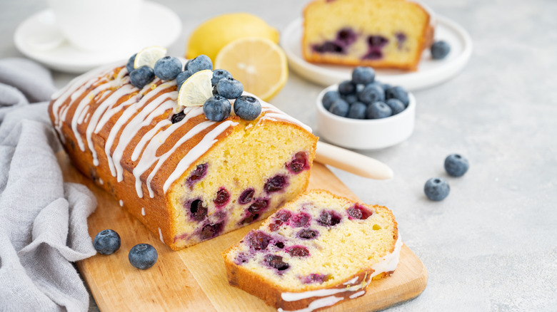 loaf of blueberry lemon pound cake with one slice o the cutting board topped with fresh blueberries and with more berries and lemons in the back