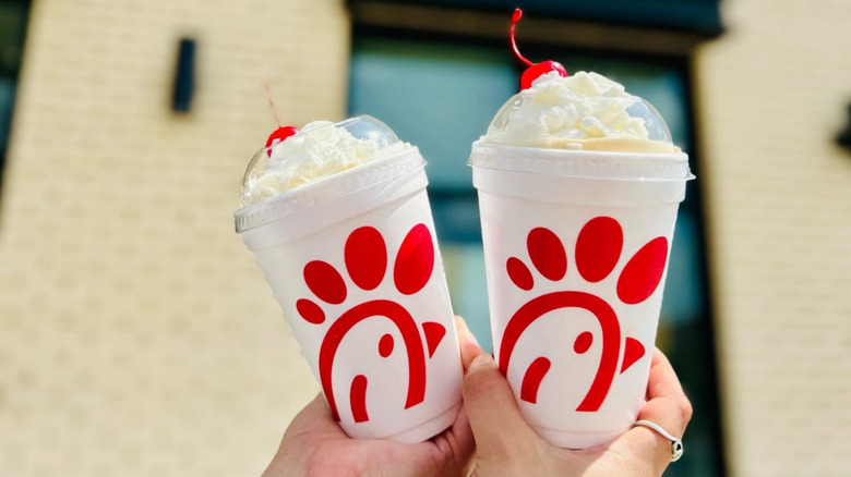 Two people holding up their peach milkshakes with cherries and whip cream up against the background of a Chick-fil-A store front.