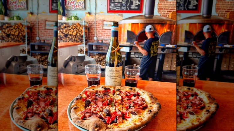 Pizza and wine set on a bar counter with a man working a pizza oven in the background