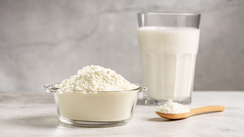 Powdered milk in glass bowl next to wooden spoon and glass of milk