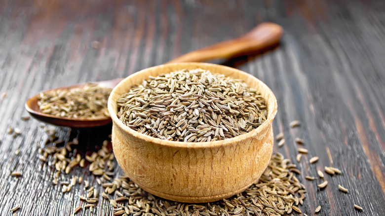 whole cumin seeds in and scattered around a light-brown bowl on a dark-brown tabletop, with a wooden spoon holding more cumin seeds visible but out of focus to the rear