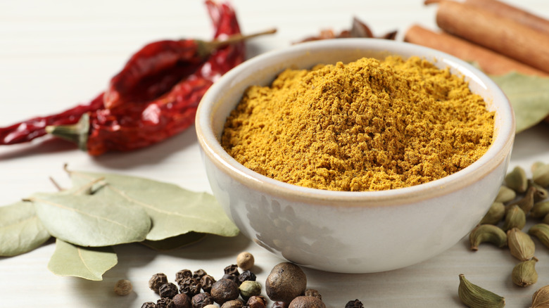 curry powder in a white bowl on a pale countertop, surrounded by other spices including cinnamon, cardamom, black pepper and chilies