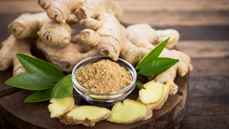 whole fresh ginger, sliced ginger, ginger leaves and a small bowl of ground dried ginger, all displayed on a wooden serving platter, on a dark-brown wooden tabletop
