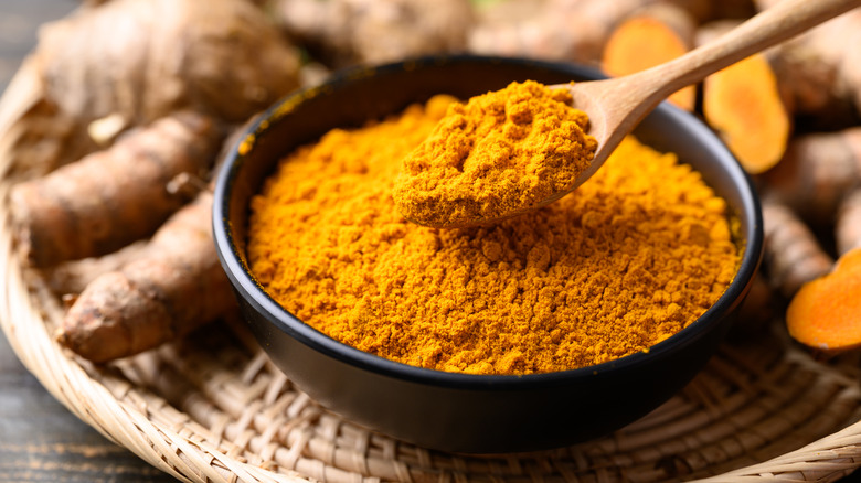 a wooden spoon scooping ground turmeric from a dark bowl on a woven mat, surrounded by fresh turmeric rhizomes