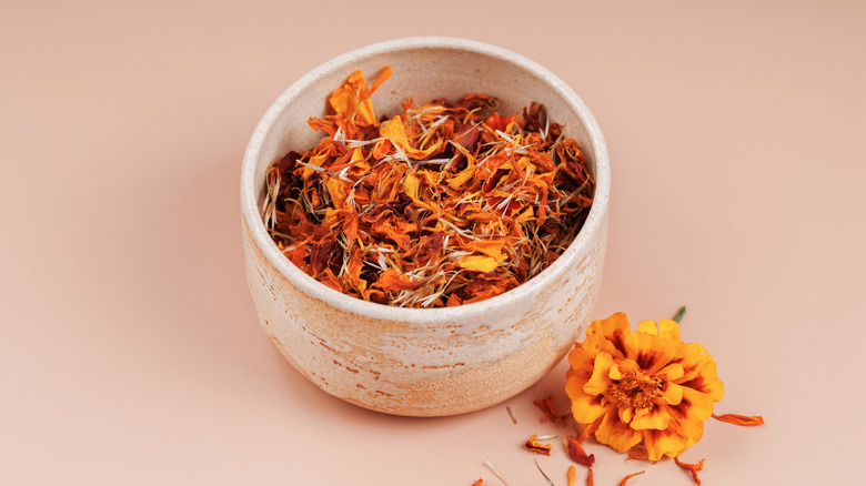 a rustic bowl filled with dried marigold leaves, on a pale-pink surface, with a fresh blossom to the front right of the bowl