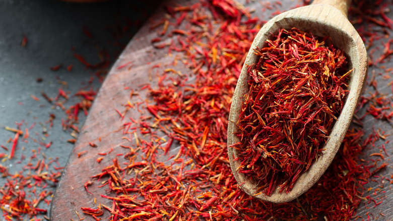 a wooden scoop of vividly colorful saffron strands resting on a cutting board, itself strewn with saffron, and resting in turn on a dark-gray stone countertop where a few strands of saffron have fallen