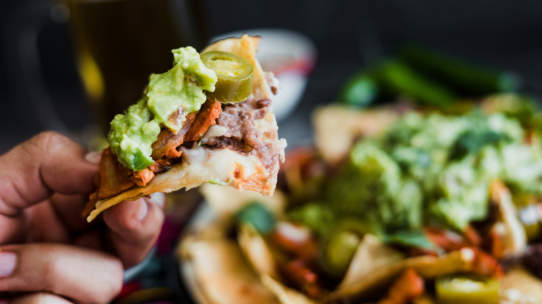 Hand holding nacho chip over plate of nachos