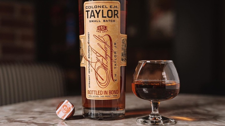 E.H. Taylor, Jr. Small Batch shown lay down on its packaging tube beside a filled whiskey glass