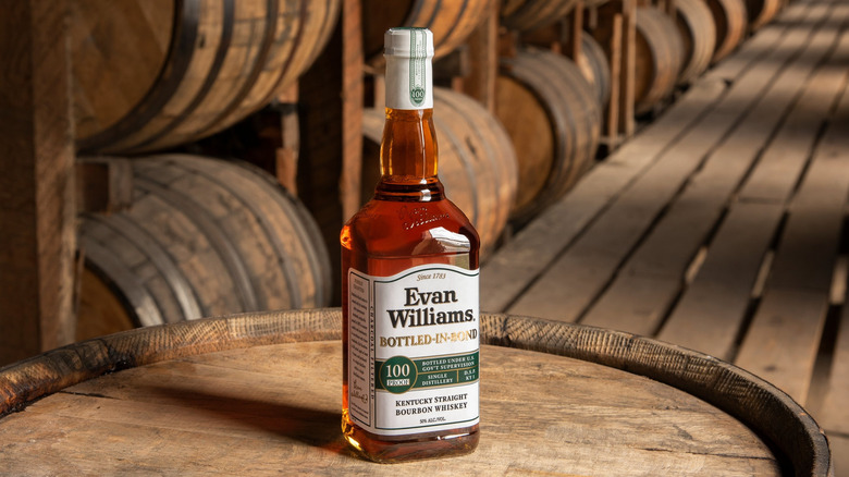 Evan Williams Bottled-in-Bond shown posed on top of a whiskey barrel with rows of barrels in the background