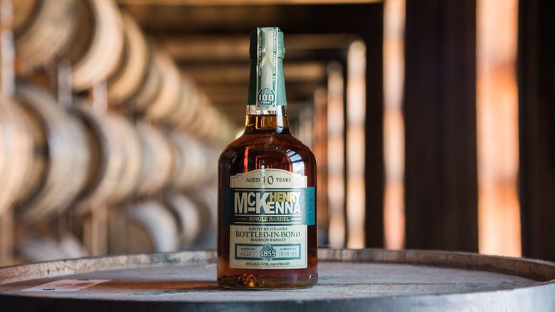 A bottle of Henry McKenna posed on top of a barrel with a blurred rickhouse shown in the background