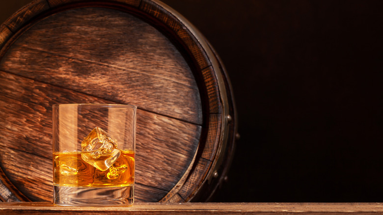 A wide shot of a filled whiskey glass with ice and a wooden barrel in the background