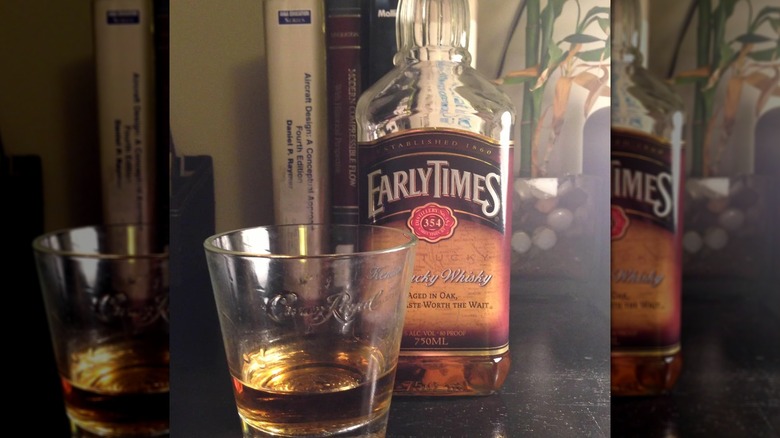 A bottle of Early Times placed on a table with a filled glass in front and books shown behind it