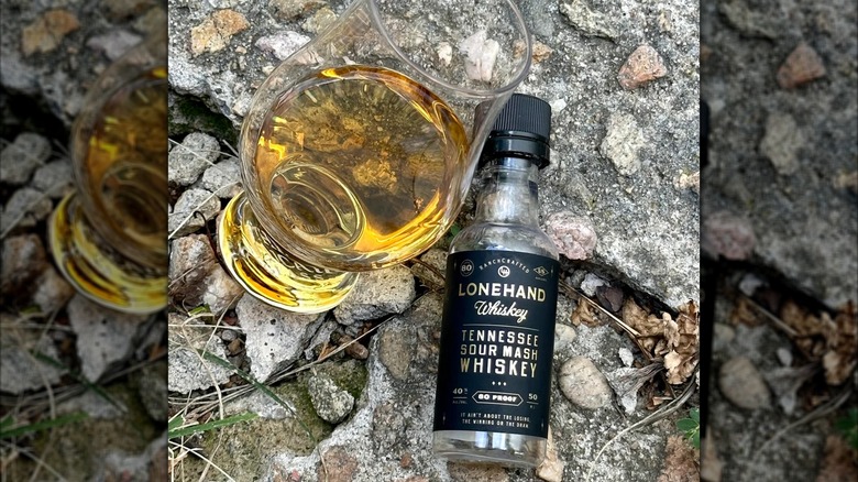 A small bottle of Lonehand whiskey placed on a rocky surface next to a filled glass