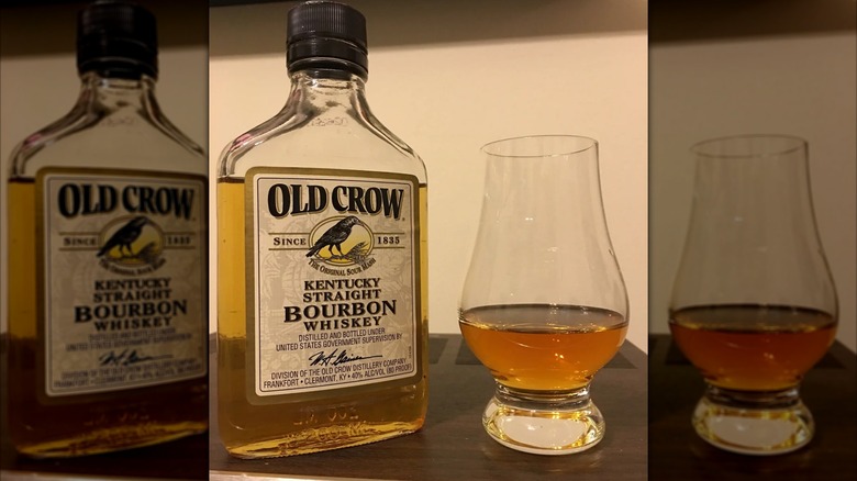 A bottle of Old Crow bourbon next to a filled glass, in front of a plain white background