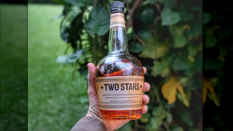 A hand holding a bottle of Two Stars bourbon up to the camera, with a blurred garden background