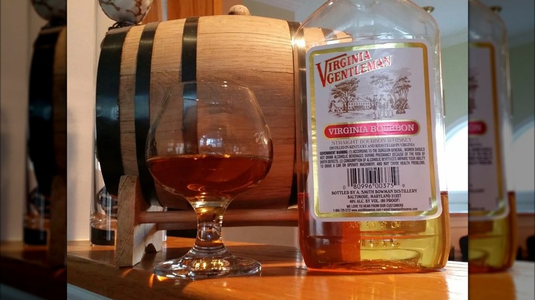 A bottle of Virginia Gentleman next to a large, filled glass, with a small barrel shown in the background