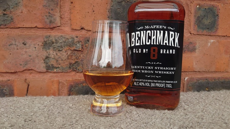 A bottle of Benchmark shown on a brick background next to a glass filled with the bourbon