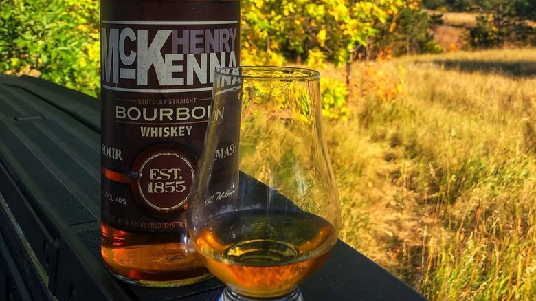 A bottle of Henry McKenna outside with a rural background and a filled class in front of it
