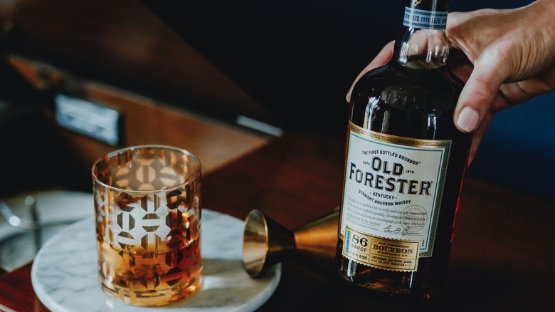 A person holding a bottle of Old Forester 86 with a glass and a measuring cup also being shown