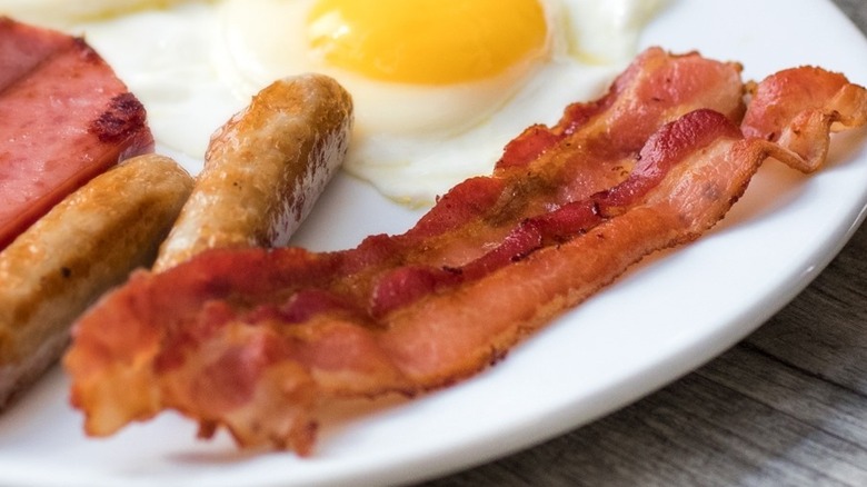 Cooked bacon next to sausage, egg and ham on plate