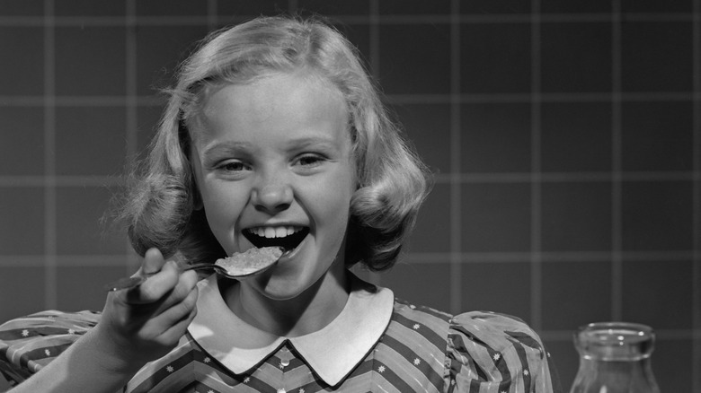 Black and white photo of girl in a dress sitting at the breakfast table putting a spoonful of cereal in her mouth