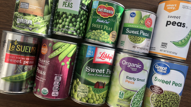 10 different brands of canned peas laying on a wooden desk.