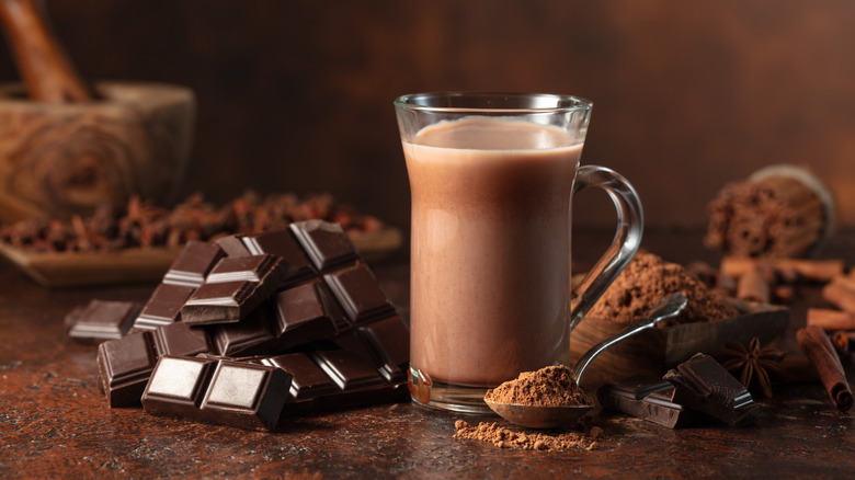 Mug of hot chocolate next to pieces of dark chocolate and spoonful of cocoa