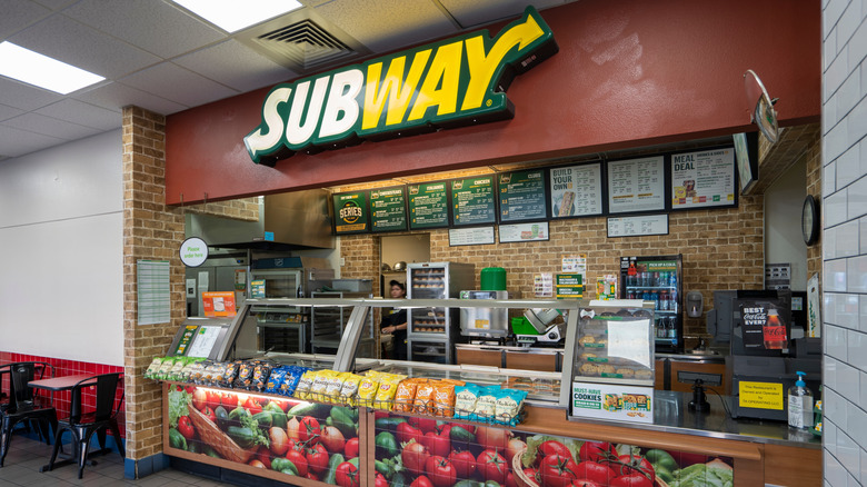 The interior of a Subway restaurant focused on the order counter/register, with menu items behind the counter and the logo above it