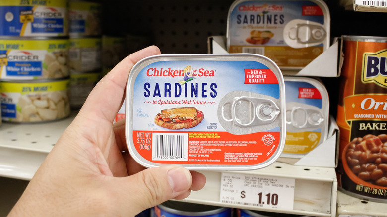A view of a hand holding a can of Chicken of the Sea sardines in front of a grocery store shelf
