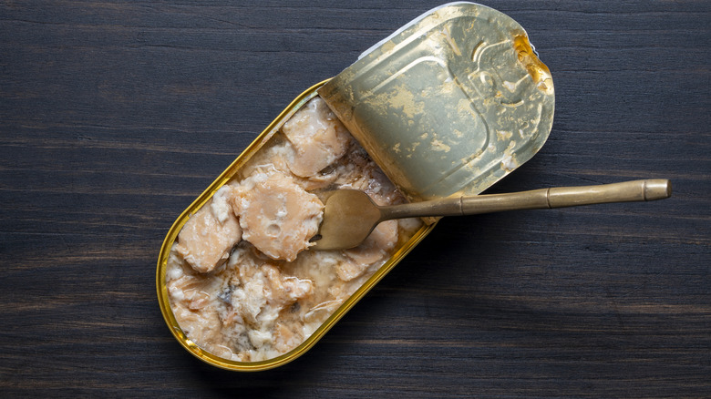 Open tin can of salmon fish fillet in its own juice and fork on dark wooden table background