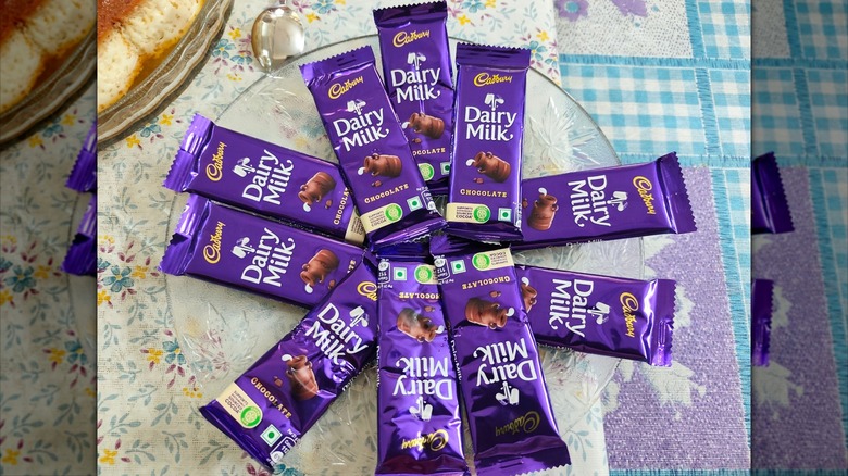 A shot of 10 Cadbury Dairy Milk bars fanned out around a circular clear glass plate on a flowery tablecloth next to blue and white checkered placemats