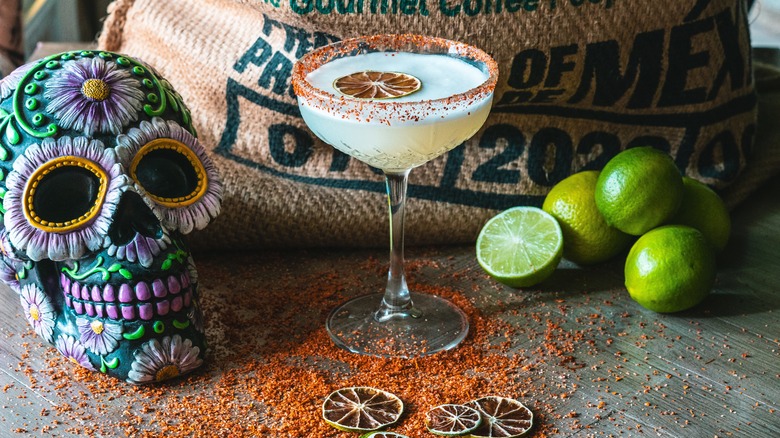 a margarita standing on top of a wooden table with some limes and tajin salt.