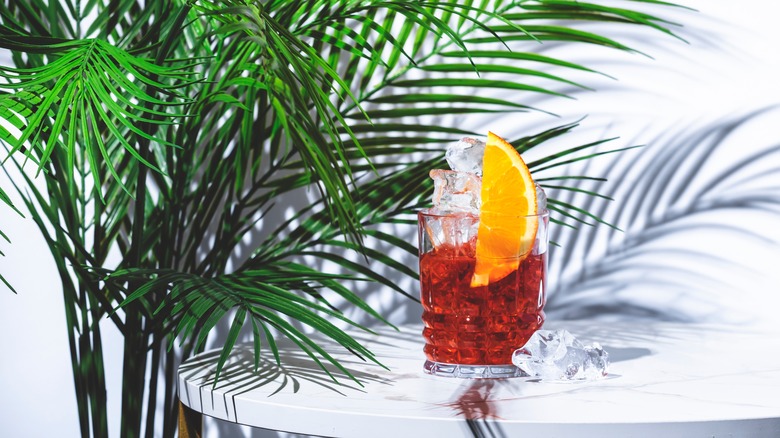 Mezcal negroni, alcoholic cocktail drink with agave spirit, red bitters, sweet vermouth, ice and orange. Marble table, white walls, palm leaves with shadow.