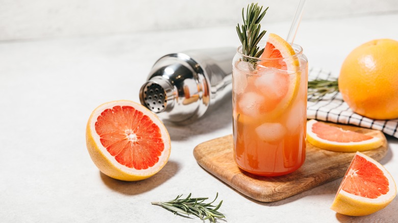 Grapefruit cocktail with rosemary garnish in a glass, served on a wooden cutting board. Vibrant, refreshing drink perfect for summer menu or cocktail recipes. Greyhound, lemonade, paloma, mocktail.