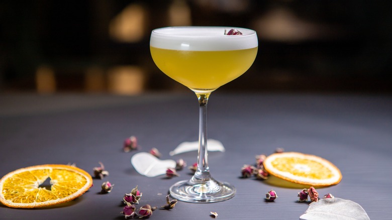 Apple Sour Gin cocktail decorated with flowers in a stemmed glass on a black table decorated with dried oranges.