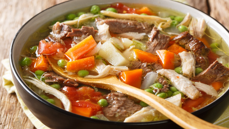 Close up of a stew with chicken, beef, potatoes, and peas with a wooden spoon sticking out of it