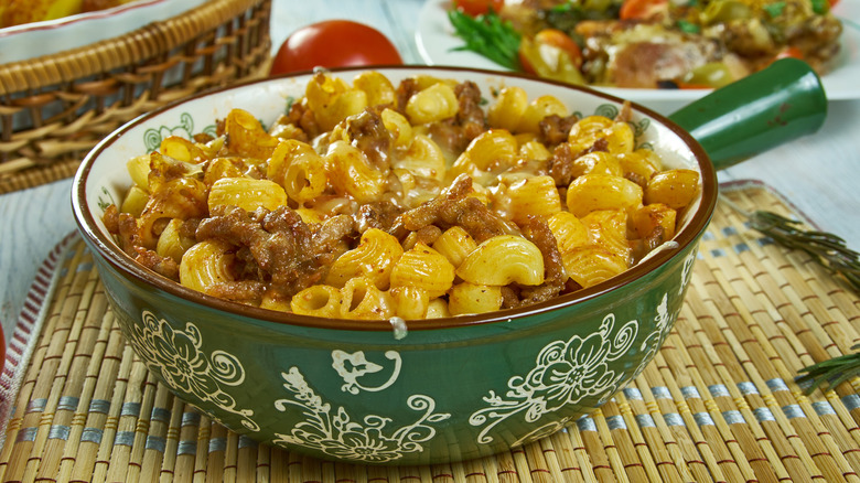 A decorative green bowl with cheesy macaroni noodles and beef on a rattan placemat